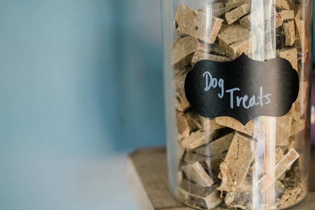 A close-up image of dry dog treats stored in a labeled glass jar on a wooden surface.